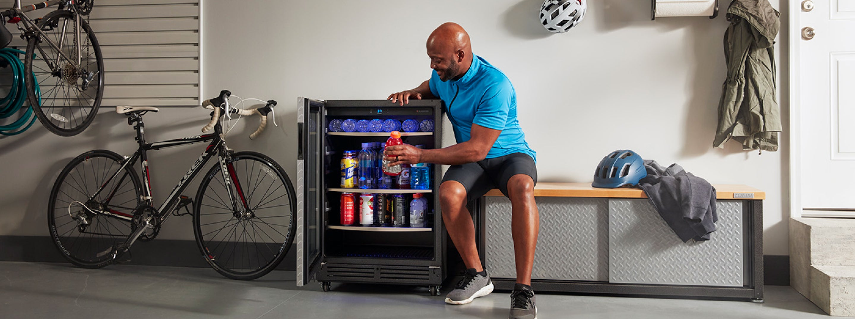 A person sitting on a Gladiator brand storage bench, retrieving an electrolyte drink from their Gladiator brand beverage refrigerator.